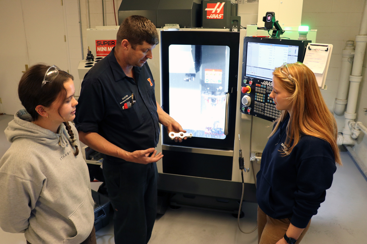 Ashley Sawa and Amber Parker listen to Dustin Burns talking the recently machined part in his hand.