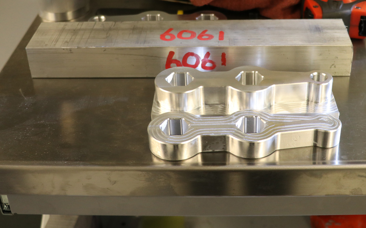 Materials on a table in the student machine shop illustrate the subtractive nature of machining from a solid block to a finished part.