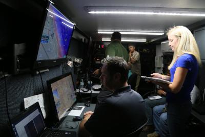 McCue, Test Director, leads a team of engineers from a control room where two remote control operators are piloting tactical drones in coordination with manned aircraft.