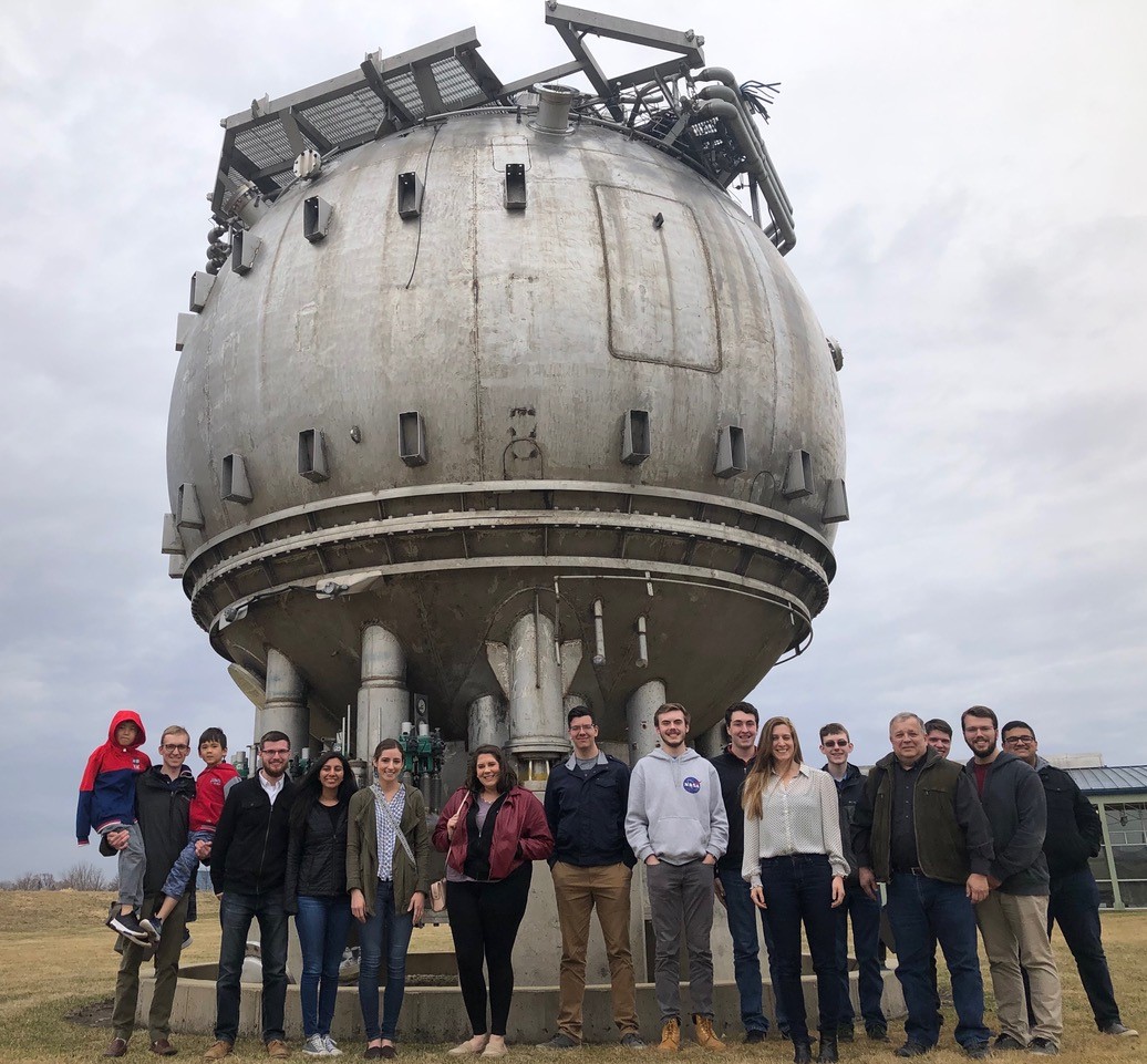 From the 2019 Fermilab visit: Standing at the foot of Fermilabâ€™s bubble chamber from left to right: Dave Stier (holding his two children), Nick Kopriva, Rachel Di Bartolomeo, Jenna Commisso, Murphy Stratton, Steve Harris, Calvin Field, Rick Eason, Stephanie Timpone, Logan Power, Michael Lembeck, Michael Harrigan, Dillon Hammond, and Avinash Rao.