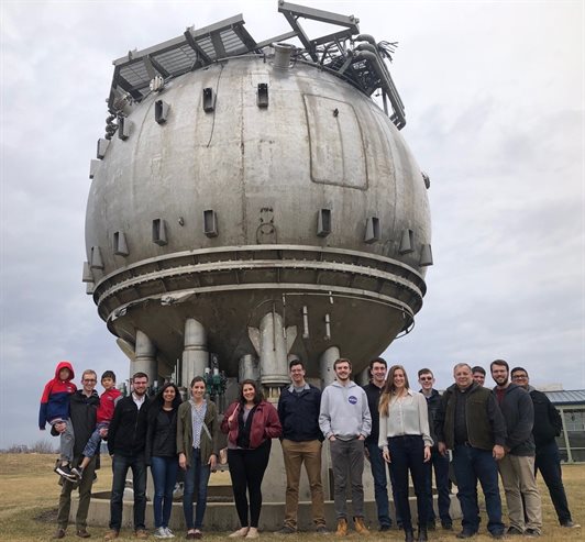 From the 2019 Fermilab visit: Standing at the foot of Fermilabâ€™s bubble chamber from left to right: Dave Stier (holding his two children), Nick Kopriva, Rachel Di Bartolomeo, Jenna Commisso, Murphy Stratton, Steve Harris, Calvin Field, Rick Eason, Stephanie Timpone, Logan Power, Michael Lembeck, Michael Harrigan, Dillon Hammond, and Avinash Rao.