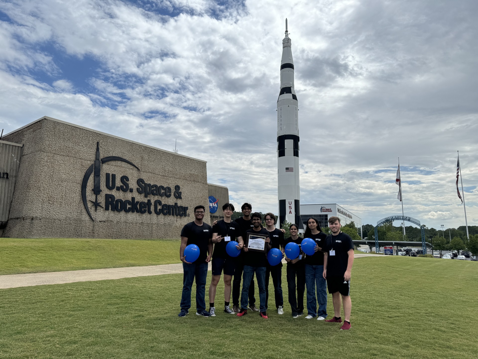 Team members with the award plaque in front of the Saturn V and US Space Rocket Center. Left to right: Sahilkrishna Vazhathodiyil, Cliff Sun, Ishaan Bansal, Shikhar Kesarwani, Adam Pawlik, Krisha Mahajan, Aparna Kamath, and Ethan Kooper.